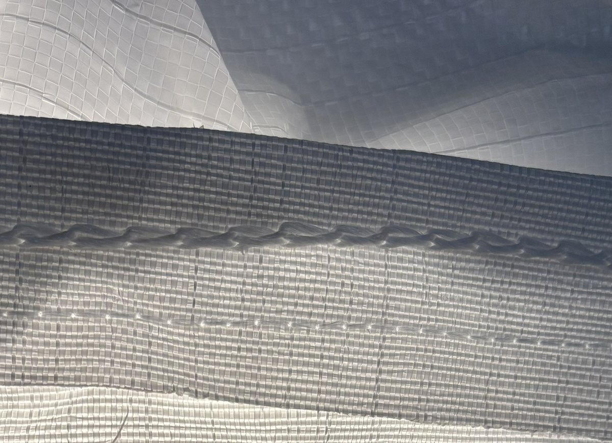 Close-up of a conventional sewn bulk-bag seam, backlit, showing the stitch line perforating the woven fabric and a loose thread end